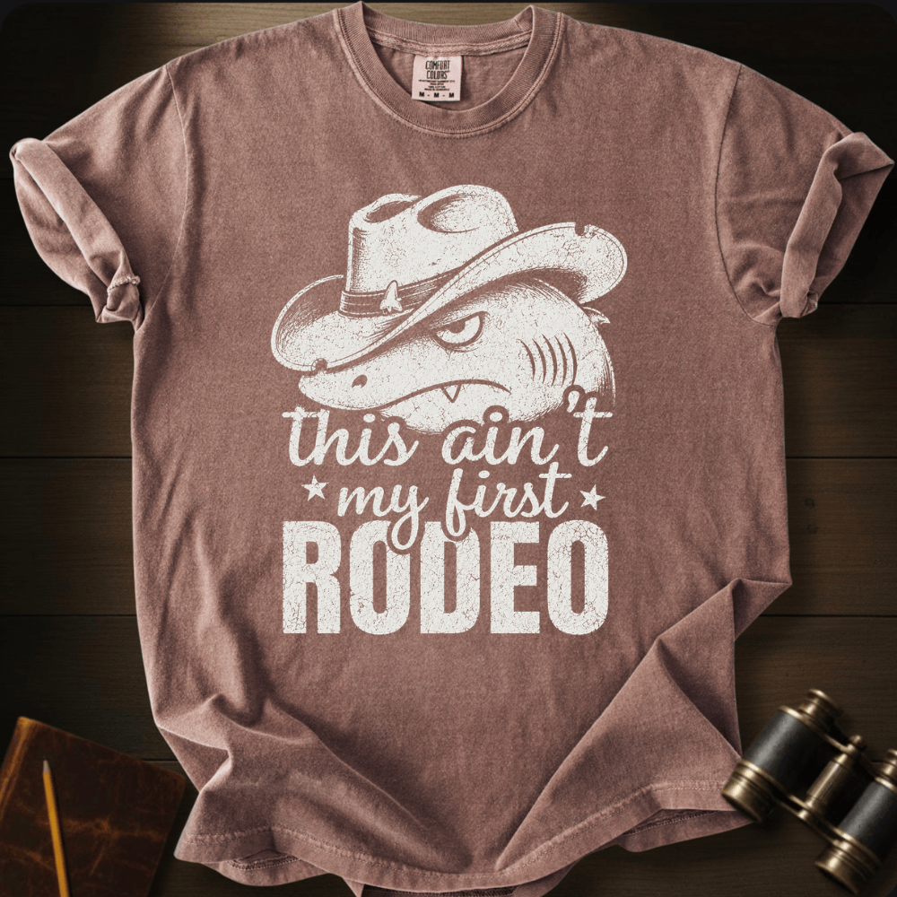 A heathered dusty rose short-sleeve t-shirt laid flat on a dark wood background, featuring a distressed white graphic of a shark wearing a cowboy hat with the text 'this ain't my first RODEO'.