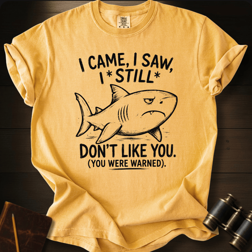A yellow graphic t-shirt laid flat on a dark wood background, featuring a black line art shark and the text 'I CAME, I SAW, I*STILL* DON'T LIKE YOU. (YOU WERE WARNED)'.