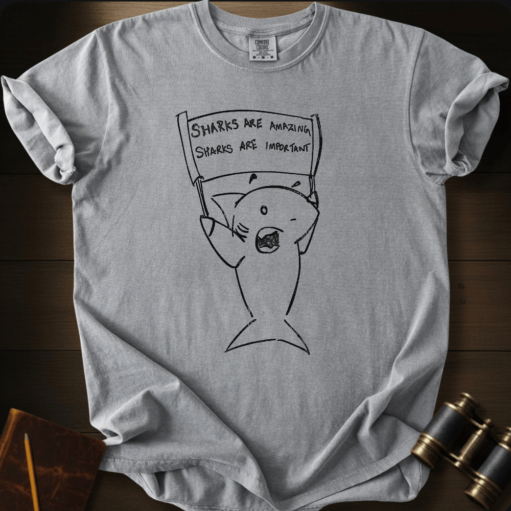 A light gray crewneck t-shirt laid flat on a dark wood background, featuring a black line art graphic of a protest shark holding a sign that reads 'SHARKS ARE AMAZING. SHARKS ARE IMPORTANT.