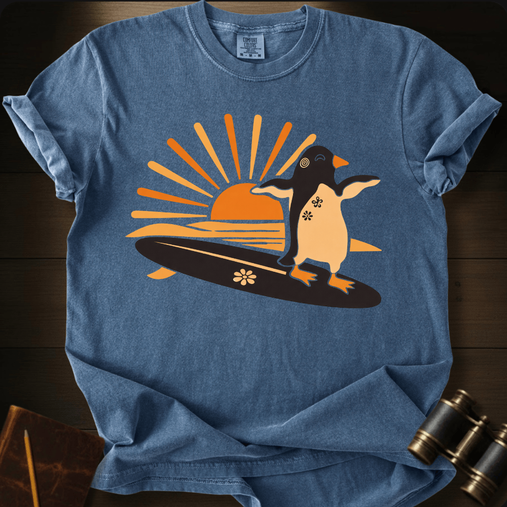 Full Sail Marine Sunset Surf penguin surfing graphic Comfort Colors t-shirt in blue jean – cute penguin on surfboard with sunset design – front view