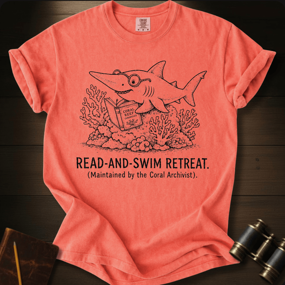 A coral pink short-sleeve t-shirt laid flat on a dark wood background, featuring a black line art graphic of a shark reading a book underwater with the text 'READ-AND-SWIM RETREAT. (Maintained by the Coral Archivist.