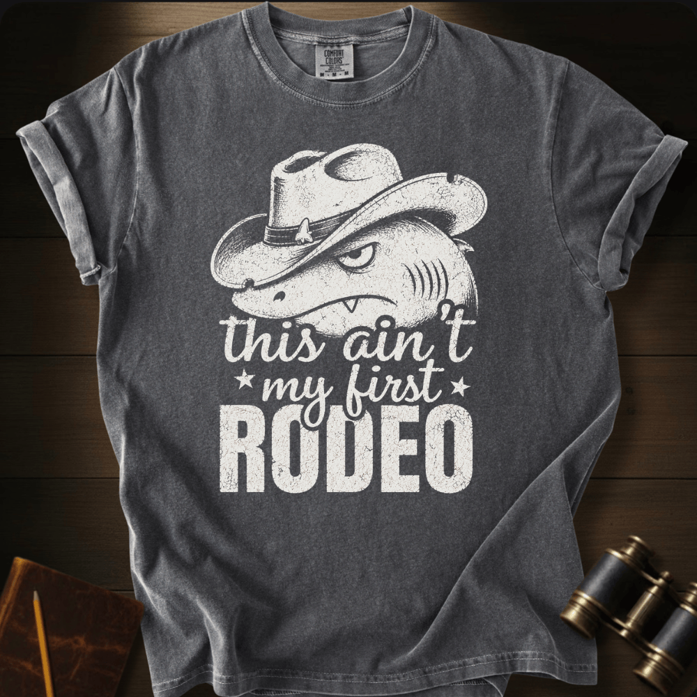 A dark gray graphic t-shirt featuring a white print of a shark wearing a cowboy hat with the text 'this ain't my first RODEO'. The t-shirt is displayed flat on a dark wood background with vintage binoculars and a notebook in the corners.
