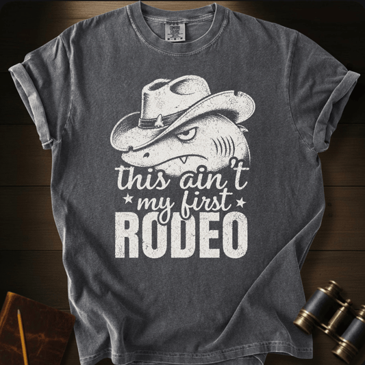 A dark gray graphic t-shirt featuring a white print of a shark wearing a cowboy hat with the text 'this ain't my first RODEO'. The t-shirt is displayed flat on a dark wood background with vintage binoculars and a notebook in the corners.