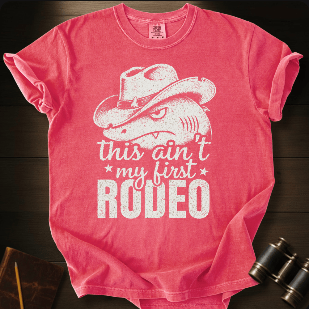 A pink short-sleeve graphic T-shirt laid flat on a dark wood surface, featuring a white distressed print of a cowboy hat and a horse head with the text 'this ain't my first RODEO.' A small neck label is visible, but no clear brand name can be read.