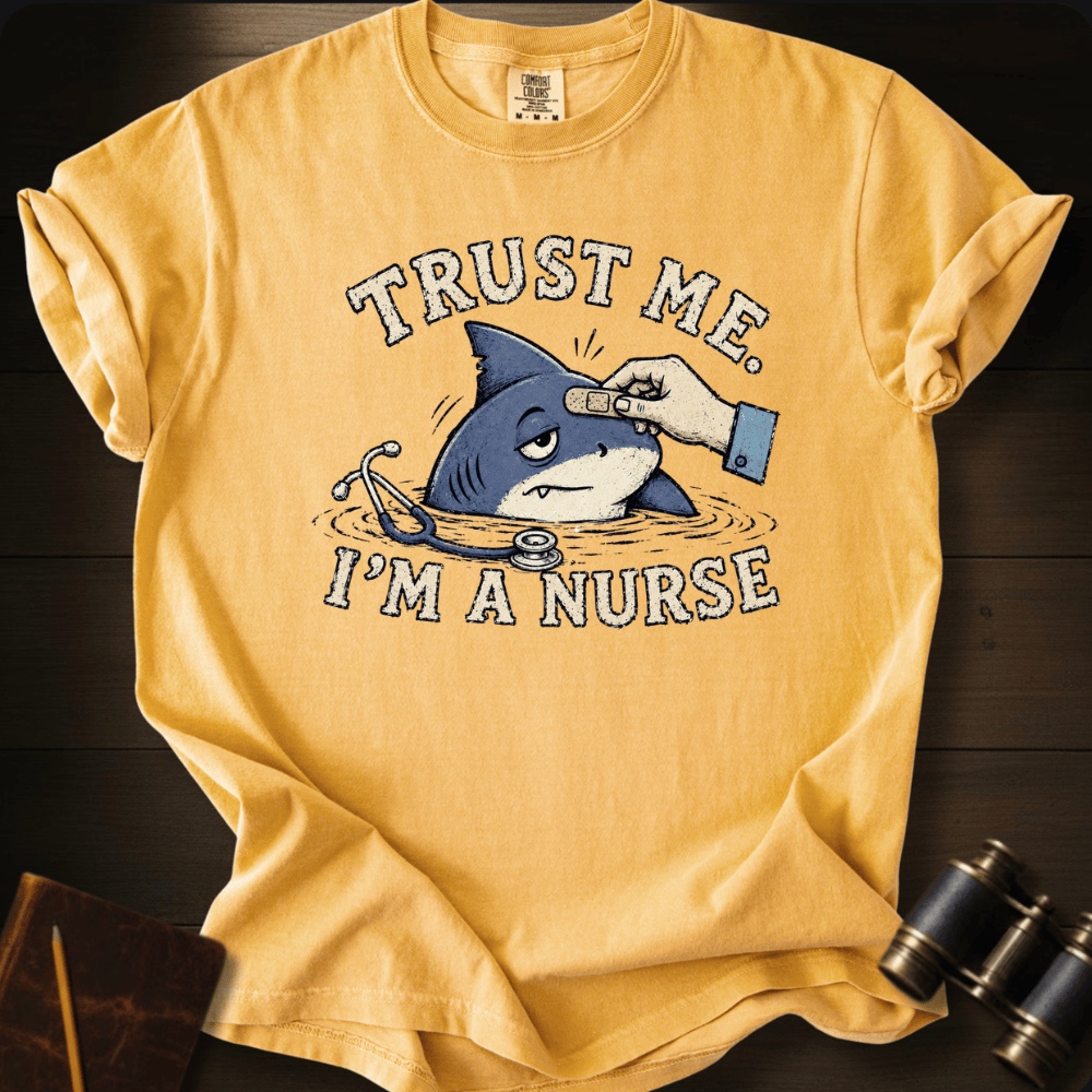 A yellow graphic t-shirt laid flat on a dark wood background, featuring a cartoon shark being checked by a nurse with the text 'TRUST ME. I'M A NURSE'.