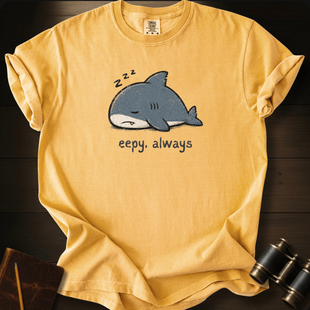 A yellow short-sleeve t-shirt laid flat on a dark wood background, featuring a cartoon sleeping shark graphic with '2zz' above it and the text 'eepy, always' below.