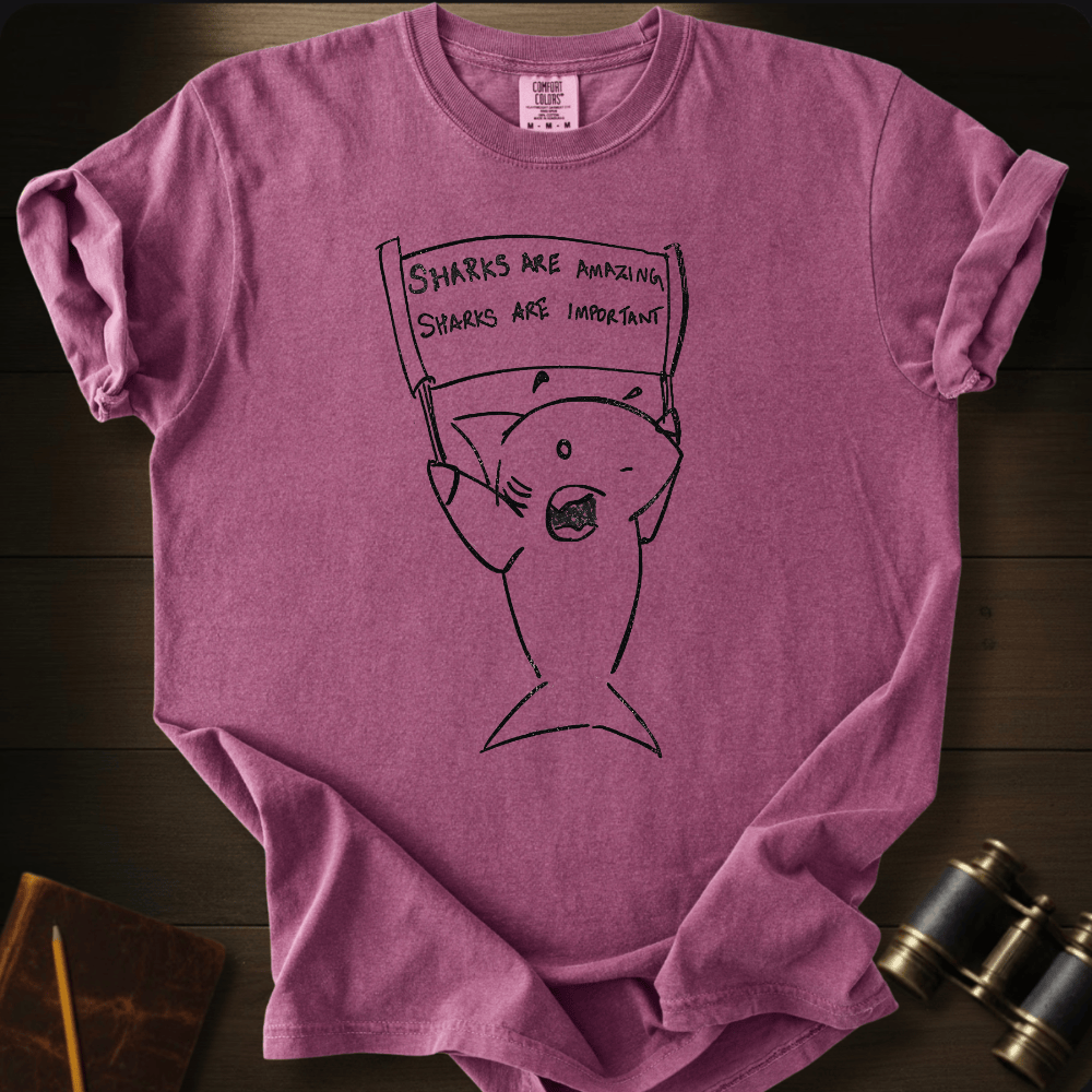 A mauve-purple short-sleeve t-shirt with a black line art graphic of a shark holding a protest sign that reads 'SHARKS ARE AMAZING SHARKS ARE IMPORTANT'. The shirt is laid flat on a dark wood background with vintage-style binoculars and a notebook in the corners.
