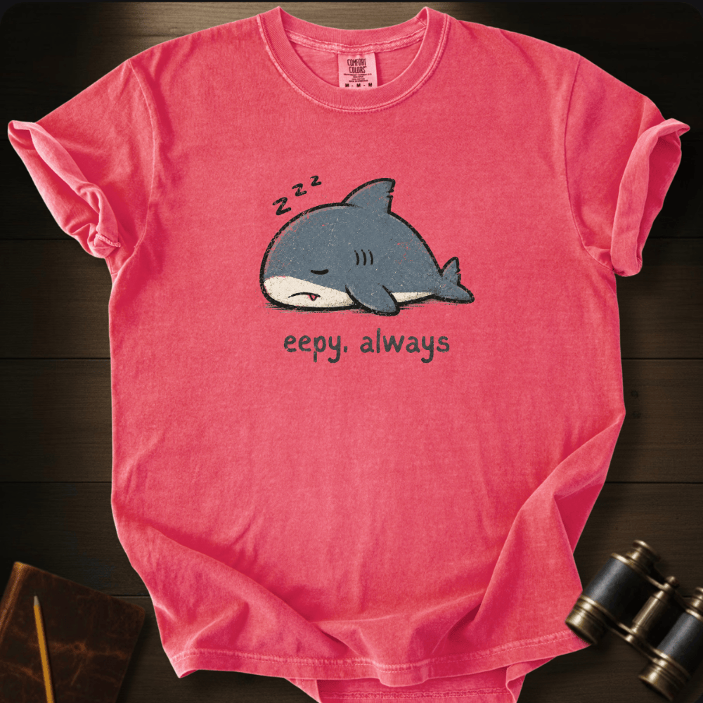 A pink short-sleeve t-shirt with a cartoon shark graphic and the text 'eepy, always' underneath, displayed flat on a dark wood background with binoculars and a notebook as props.