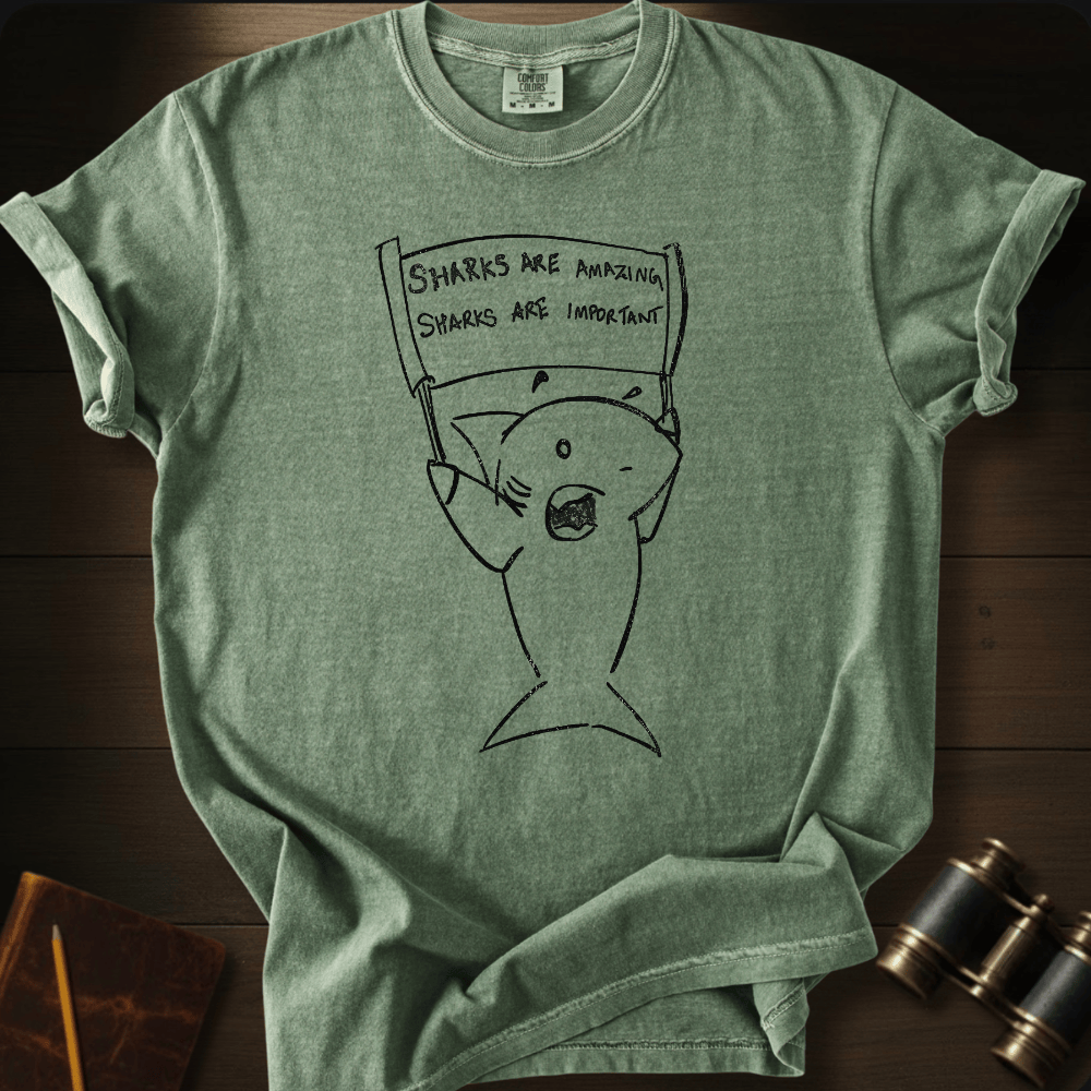 A heather sage green t-shirt with a black line art graphic of a shark holding a protest sign that reads 'SHARKS ARE AMAZING. SHARKS ARE IMPORTANT!'.