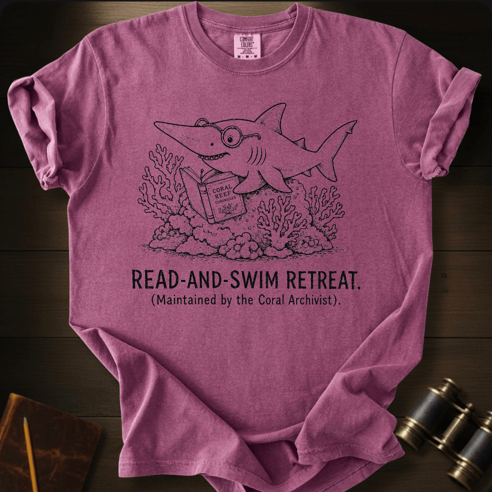 A heathered mauve pink short-sleeve t-shirt with a black line art graphic of a shark reading under coral, with the text 'READ-AND-SWIM RETREAT. (Maintained by the Coral Archivist.
