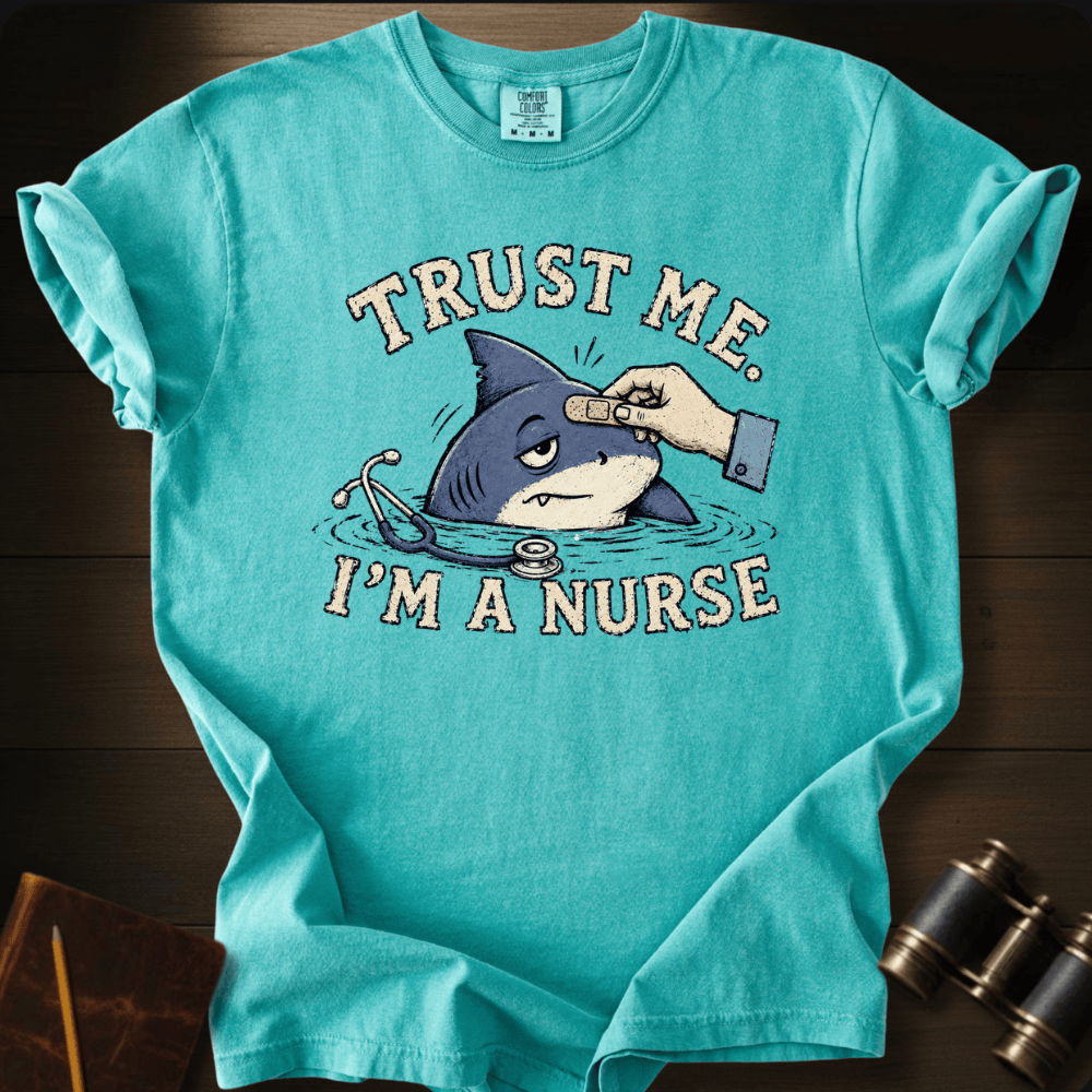 A turquoise graphic t-shirt laid flat on a dark wood background, featuring a cartoon shark in a pool with a bandage on its head and the text 'TRUST ME. I'M A NURSE'.