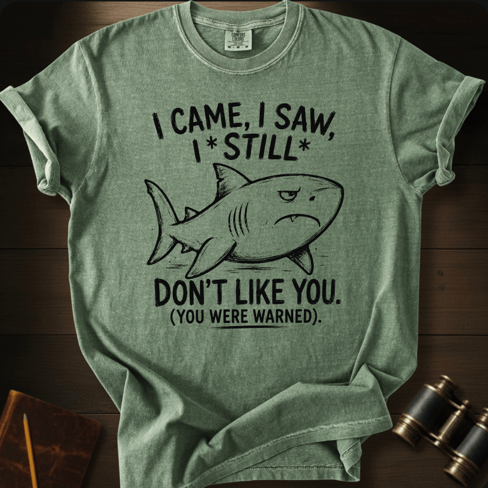 A heather sage green graphic t-shirt laid flat on a dark wood background. The shirt features a black line art illustration of a shark and the text 'I CAME, I SAW, I * STILL * DON'T LIKE YOU.