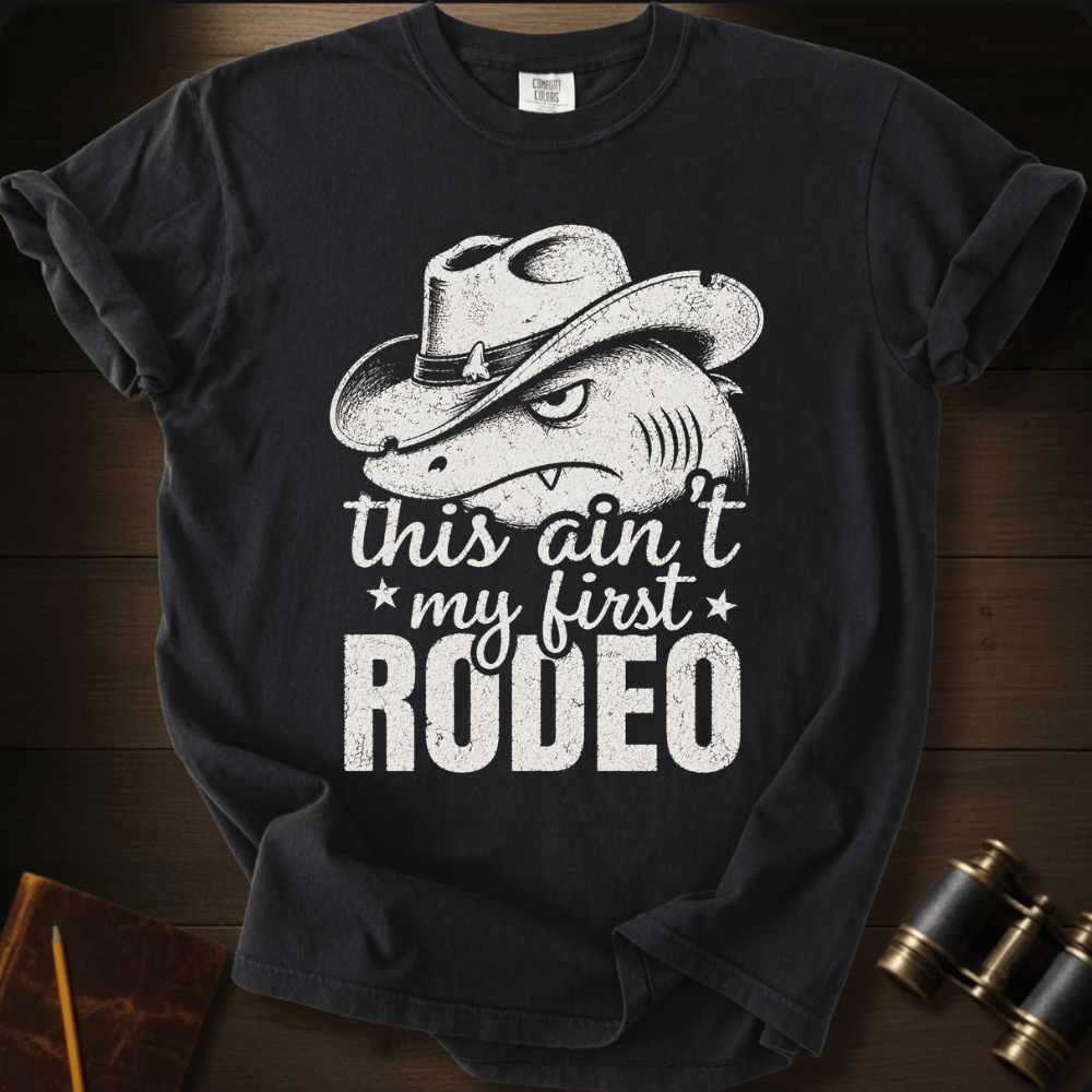 A black short-sleeve graphic T-shirt laid flat on a dark wood surface, featuring a distressed white print of a shark wearing a cowboy hat with the text 'this ain't my first RODEO.' Small props, including a notebook and binoculars, are partially visible around the shirt.