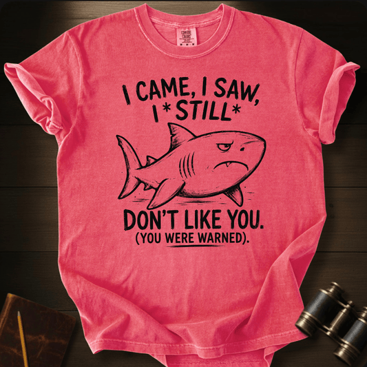 A pink short-sleeve graphic T-shirt laid flat on a dark wood surface, featuring a black shark illustration and the text 'I CAME, I SAW, I * STILL * DON'T LIKE YOU. (YOU WERE WARNED).