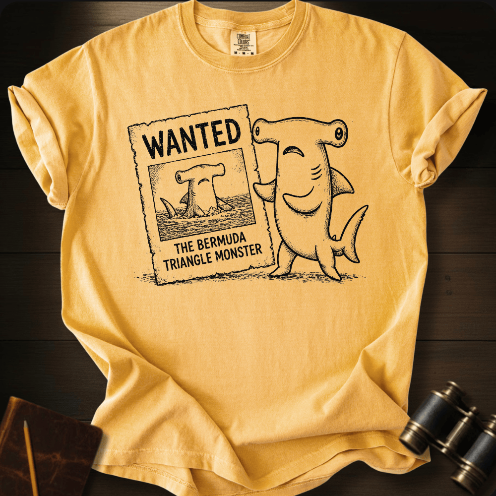 Mustard Comfort Colors tee with a cartoon hammerhead shark holding a 'Wanted: The Bermuda Triangle Monster' poster.