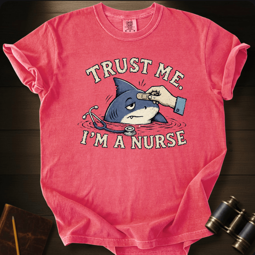 A pink short-sleeve graphic T-shirt laid flat on a dark wood surface, featuring a cartoon shark with a stethoscope and a hand placing a bandage on the shark, with the text 'TRUST ME. I’M A NURSE.
