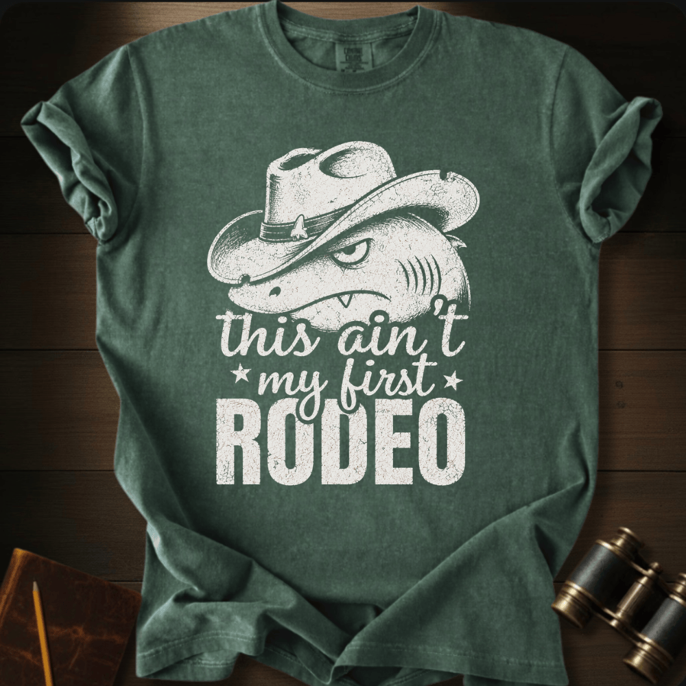 A dark green heathered t-shirt with a distressed white graphic of a shark wearing a cowboy hat and the text 'this ain't my first RODEO', displayed flat on a dark wood background with vintage binoculars and a notebook as props.