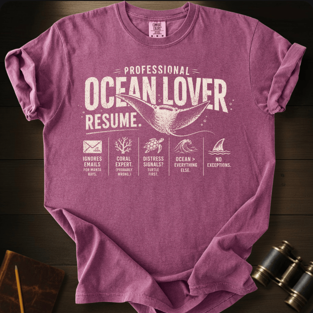 A heathered purple short-sleeve t-shirt with a white graphic print reading 'PROFESSIONAL OCEAN LOVER RESUME.' laid flat on a dark wood background, with small nautical-themed icons and a pair of binoculars partially visible in the corner.