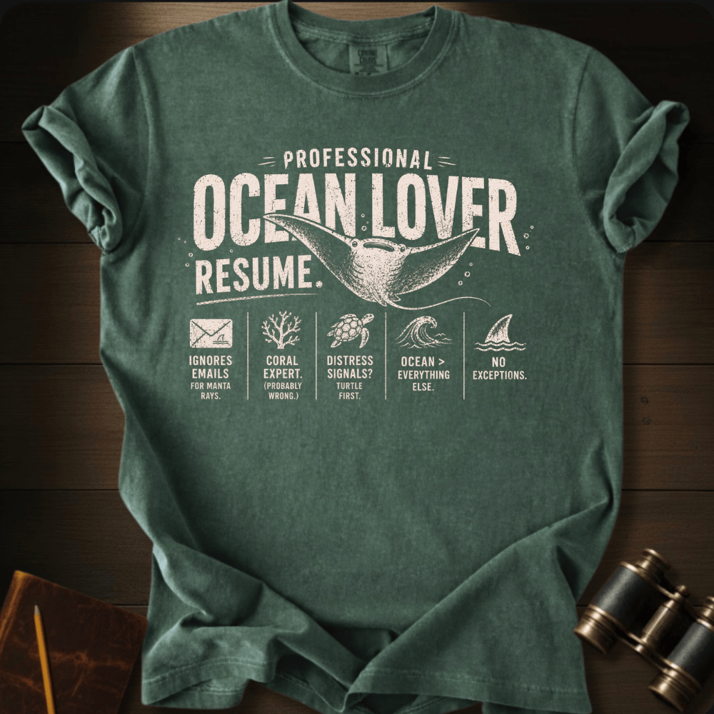 A hemp graphic t-shirt laid flat on a dark wood background, featuring a white distressed print that reads 'PROFESSIONAL OCEAN LOVER RESUME.' with sea-themed icons and a whale tail graphic.