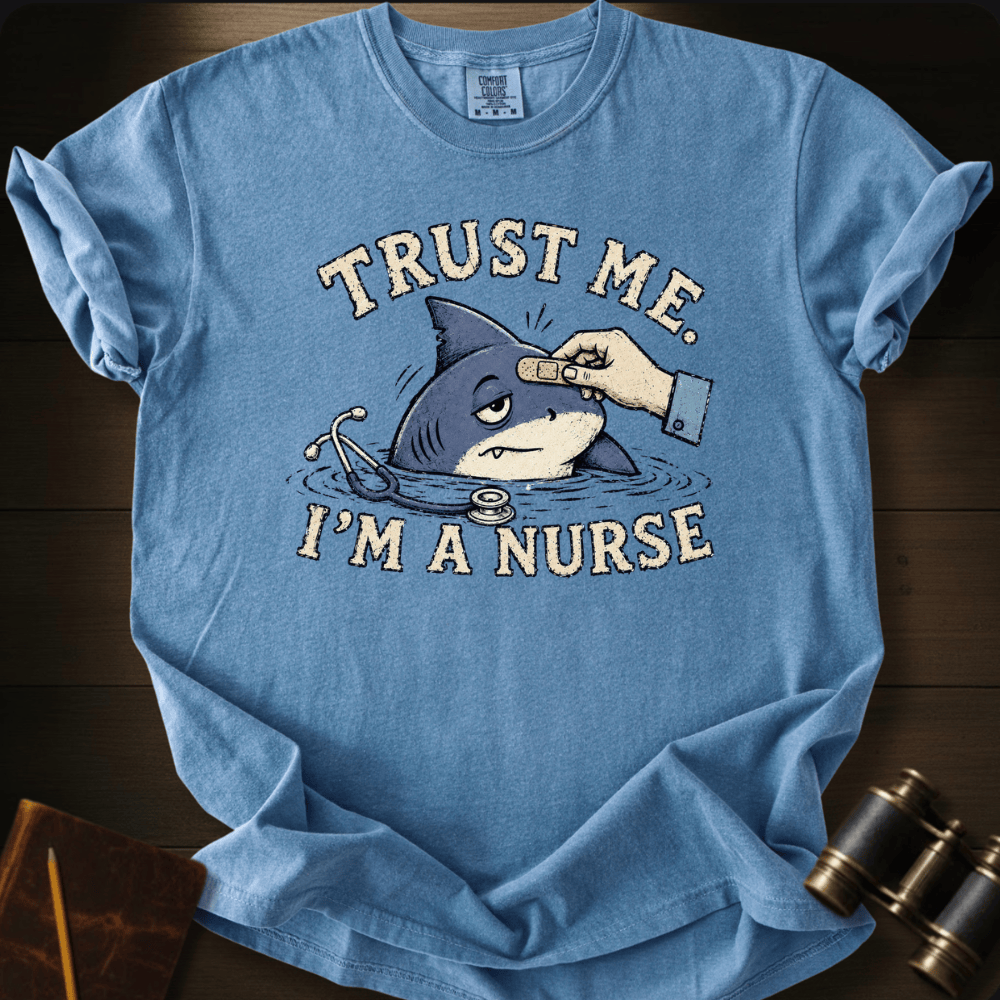 A light blue graphic t-shirt laid flat on a dark wood background. The shirt features a cartoon illustration of a shark with a bandage on its head and a stethoscope, with the text 'TRUST ME.