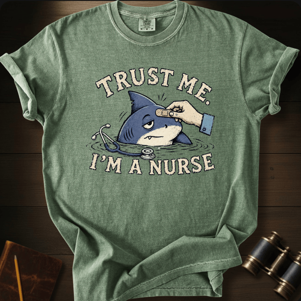 A heather sage green graphic t-shirt laid flat on a dark wood background. The shirt features a cartoon illustration of a shark with a bandage on its head and a stethoscope, with the text 'TRUST ME.
