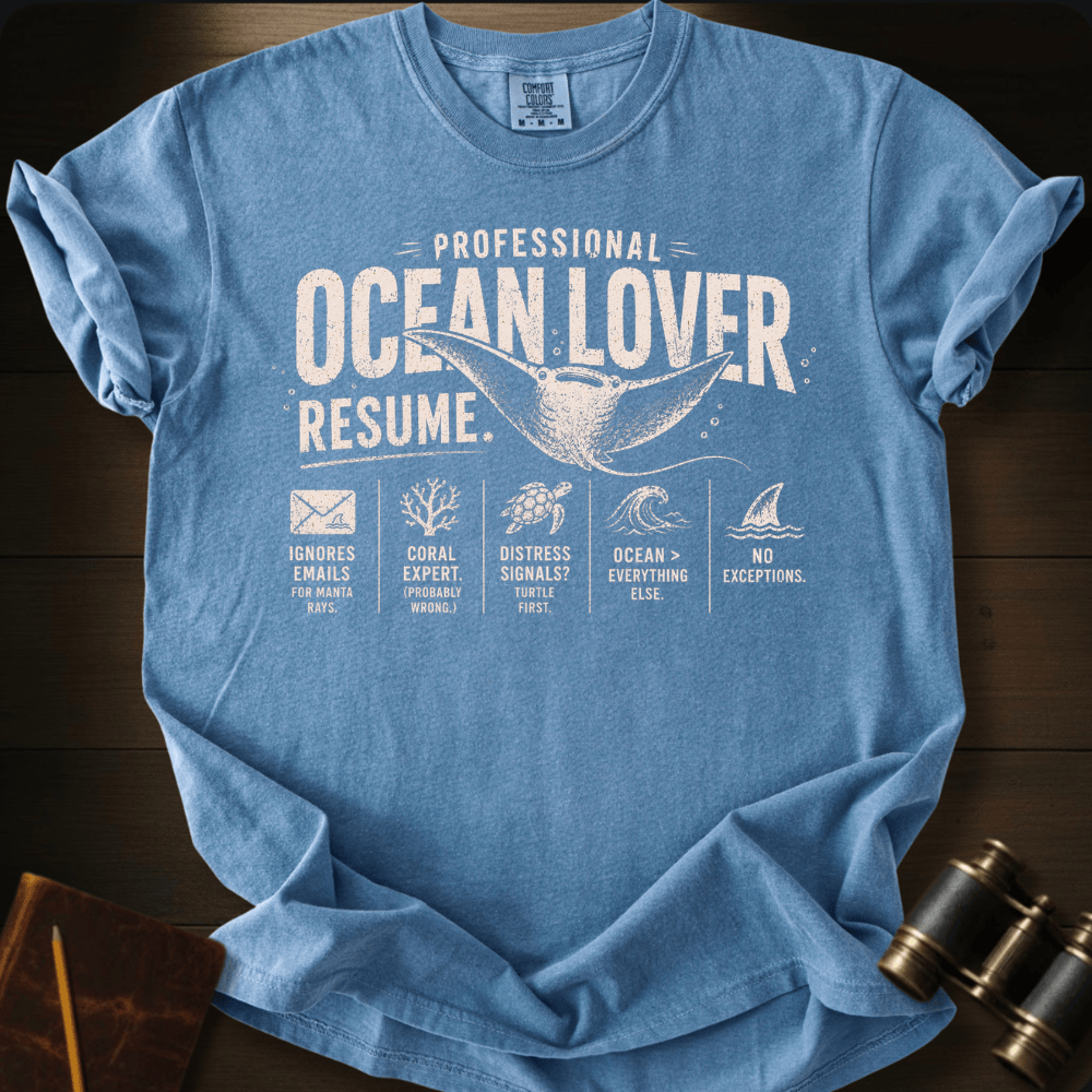 A light blue graphic t-shirt laid flat on a dark wood background, featuring a white printed resume-style design with a whale, waves, and fish icons under the text 'PROFESSIONAL OCEAN LOVER RESUME.'.