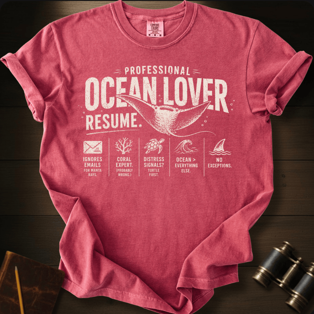 A crimson pink graphic t-shirt laid flat on a dark wood background, styled with a notebook, pencil, and binoculars. The shirt features a white distressed print that reads 'PROFESSIONAL OCEAN LOVER RESUME.