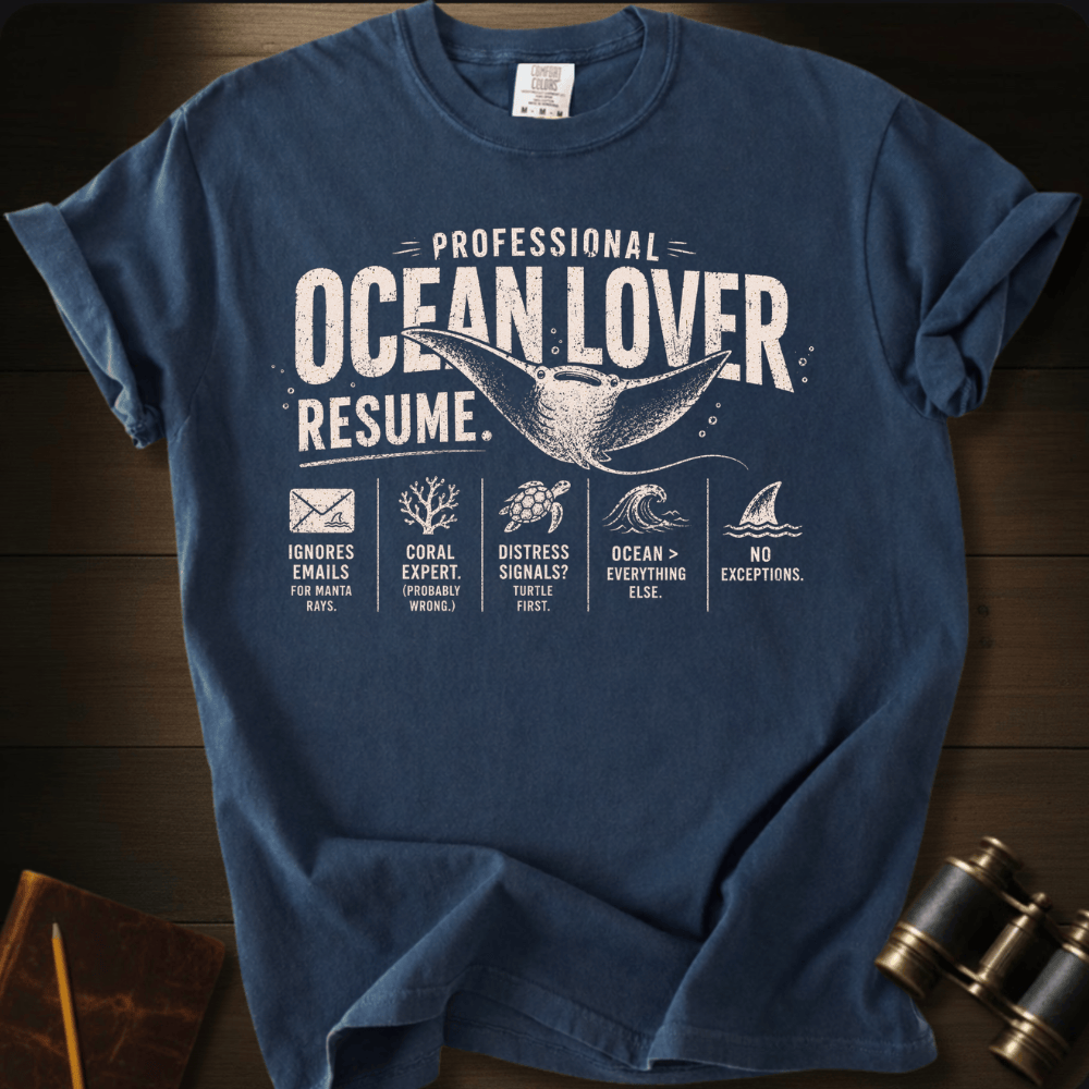 A navy blue graphic t-shirt laid flat on a dark wood background, featuring a white distressed print that reads 'PROFESSIONAL OCEAN LOVER RESUME.' with a whale tail illustration and small ocean-themed icons and text.