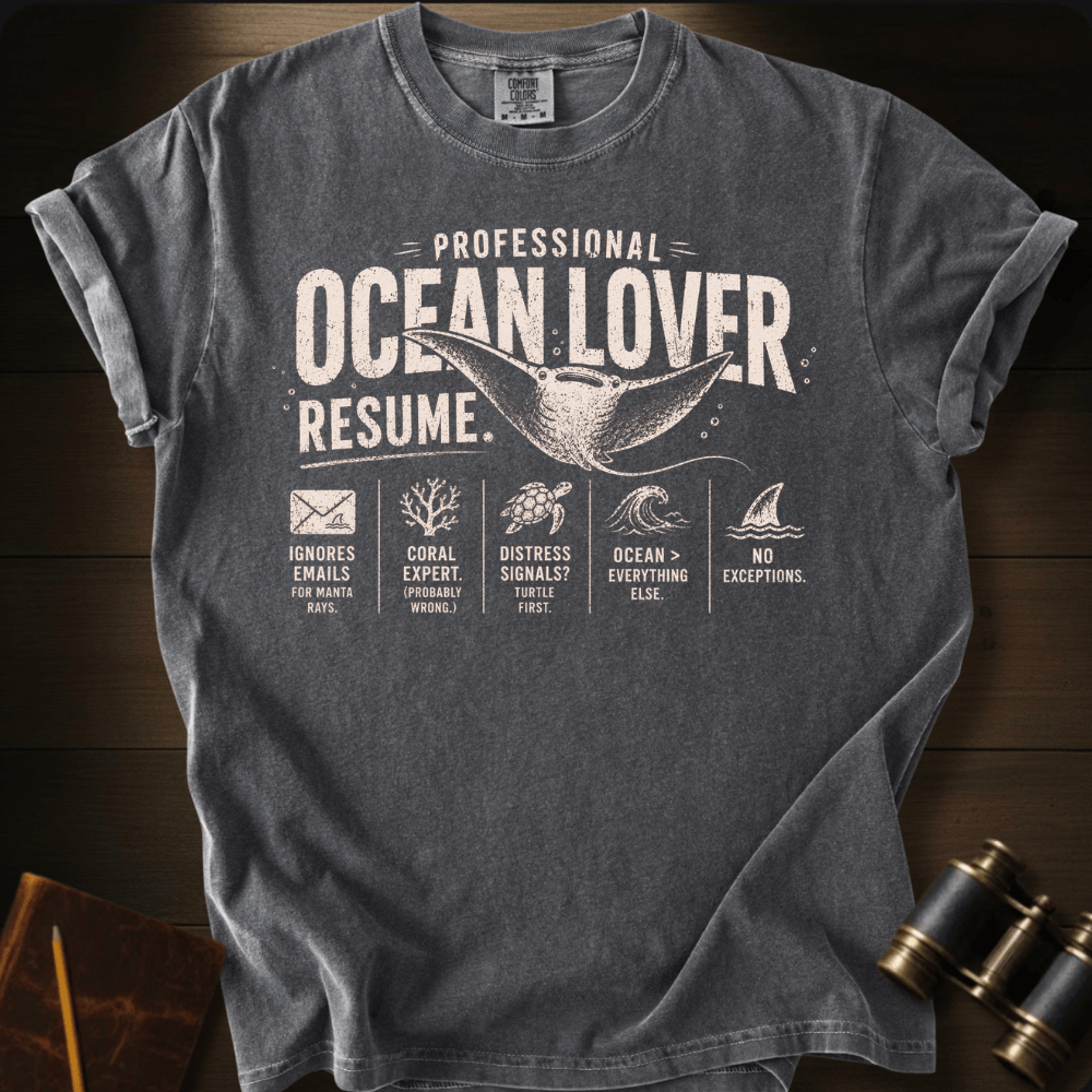 A pepper graphic t-shirt laid flat on a dark wood background, styled with a vintage explorer theme including binoculars and a notebook. The t-shirt features a humorous resume design for an 'Ocean Lover' with illustrations of a whale, sea turtle, shark, coral, and waves.