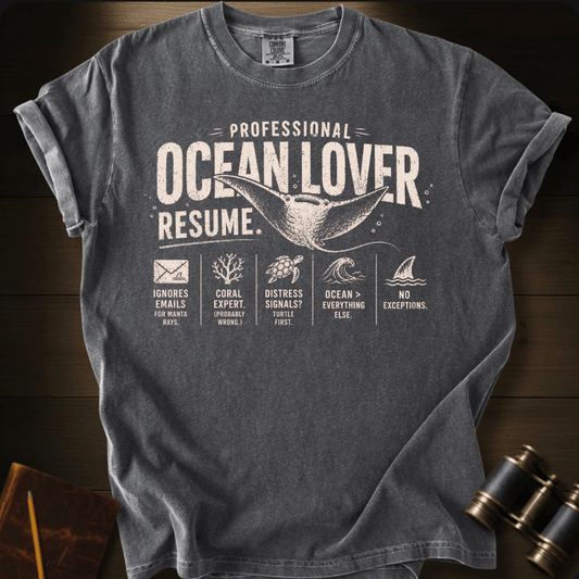 A pepper graphic t-shirt laid flat on a dark wood background, styled with a vintage explorer theme including binoculars and a notebook. The t-shirt features a humorous resume design for an 'Ocean Lover' with illustrations of a whale, sea turtle, shark, coral, and waves.