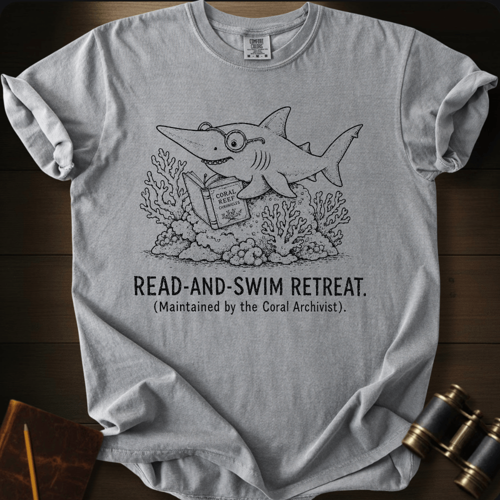 A light gray short-sleeve t-shirt laid flat on a dark wood background, featuring a black line art graphic of a shark reading a book underwater with the text 'READ-AND-SWIM RETREAT. (Maintained by the Coral Archivist.