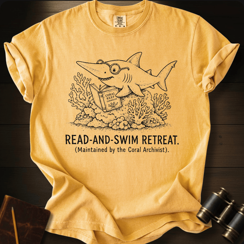 A yellow graphic t-shirt laid flat on a dark wood background, featuring a black line art illustration of a shark reading a book with the text 'READ-AND-SWIM RETREAT. (Maintained by the Coral Archivist).