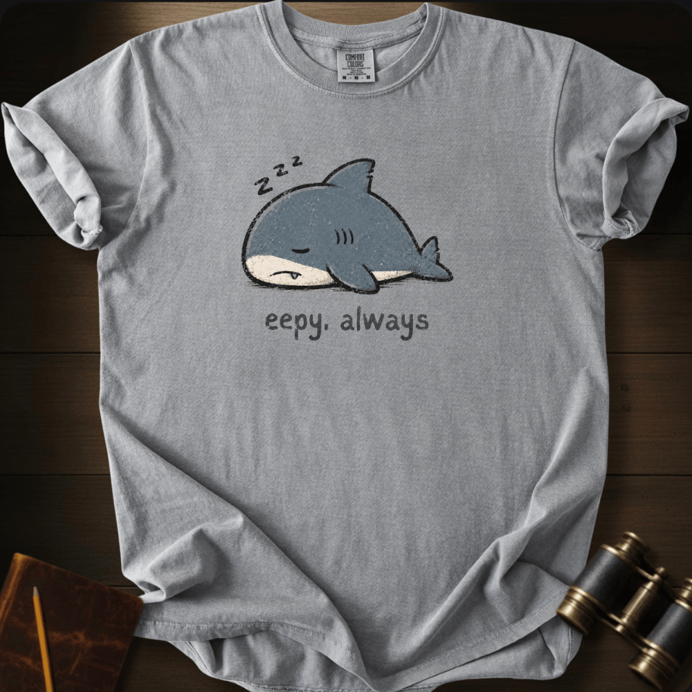 A light gray crewneck t-shirt laid flat on a dark wood background, featuring a cartoon sleeping shark graphic with 'zzz' above it and the text 'eepy. always' below.
