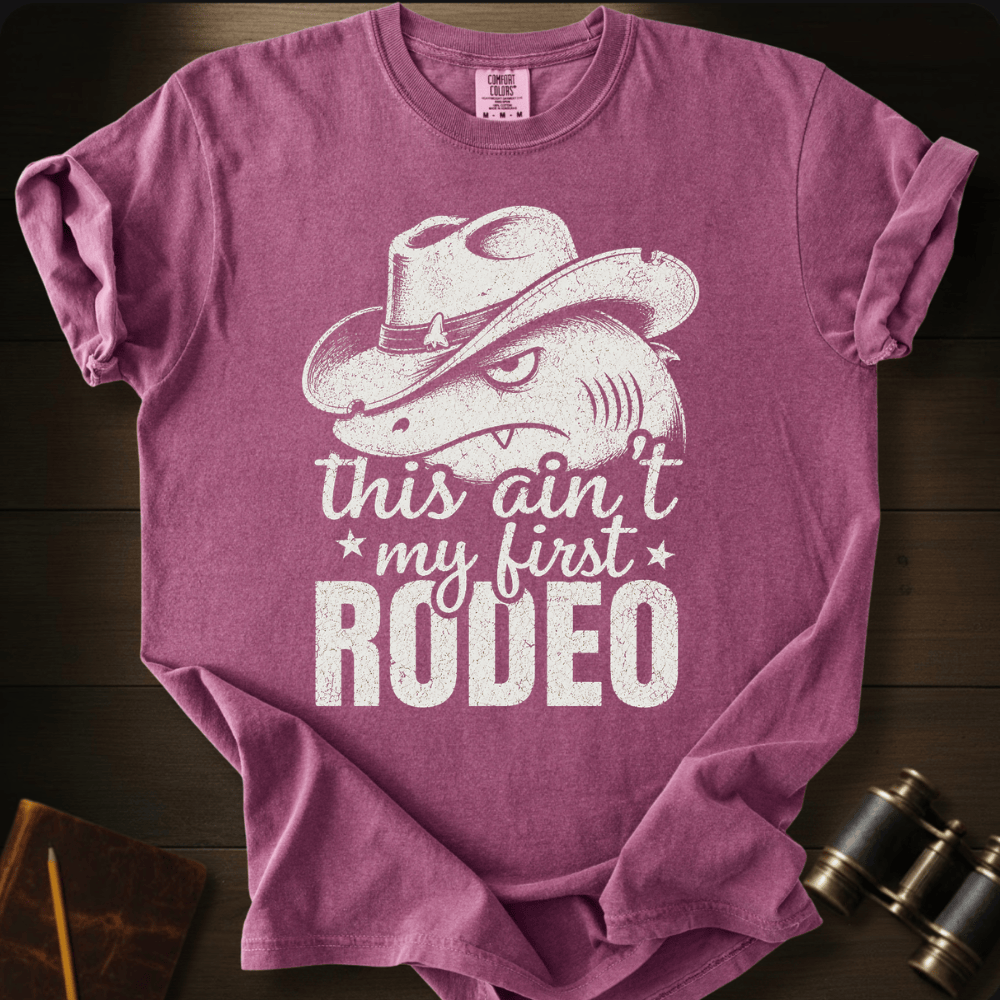 A heathered purple short-sleeve t-shirt with a distressed white graphic of a shark wearing a cowboy hat and the text 'this ain't my first RODEO', laid flat on a dark wood background with vintage binoculars and a notebook as props.