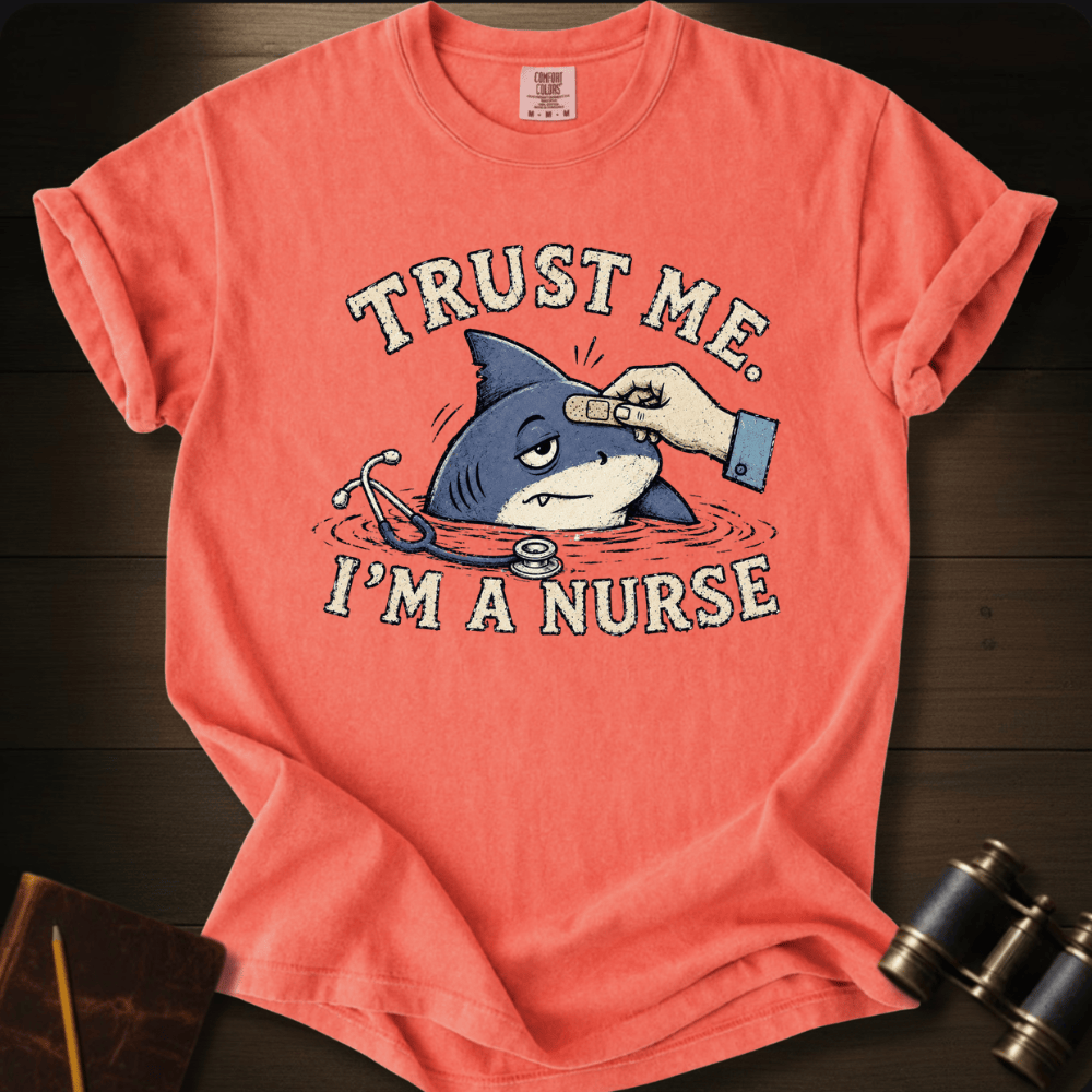 A coral pink graphic t-shirt laid flat on a dark wood background. The shirt features a cartoon illustration of a shark in a stethoscope with a hand giving an injection, and the text 'TRUST ME.
