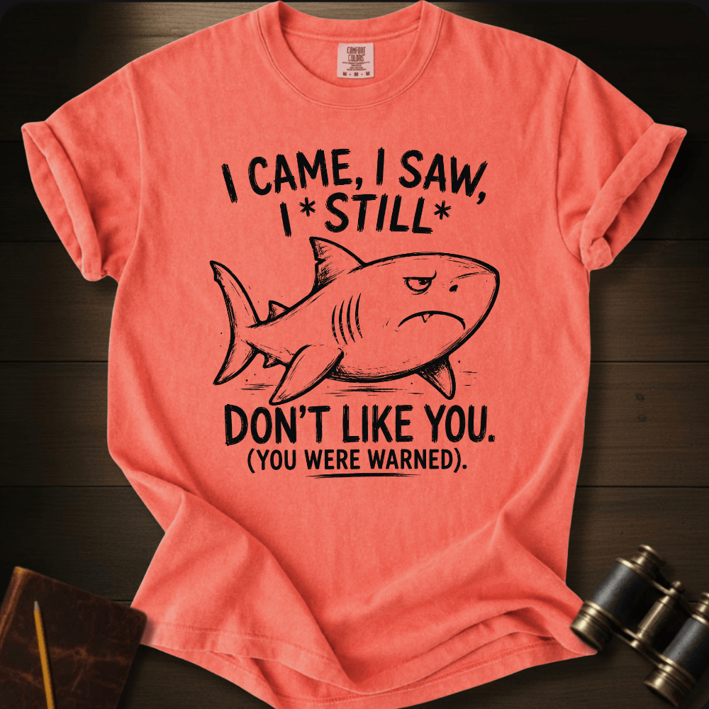 A coral pink graphic t-shirt laid flat on a dark wood background, featuring a black line art shark and the text 'I CAME, I SAW, I * STILL * DON'T LIKE YOU. (YOU WERE WARNED).