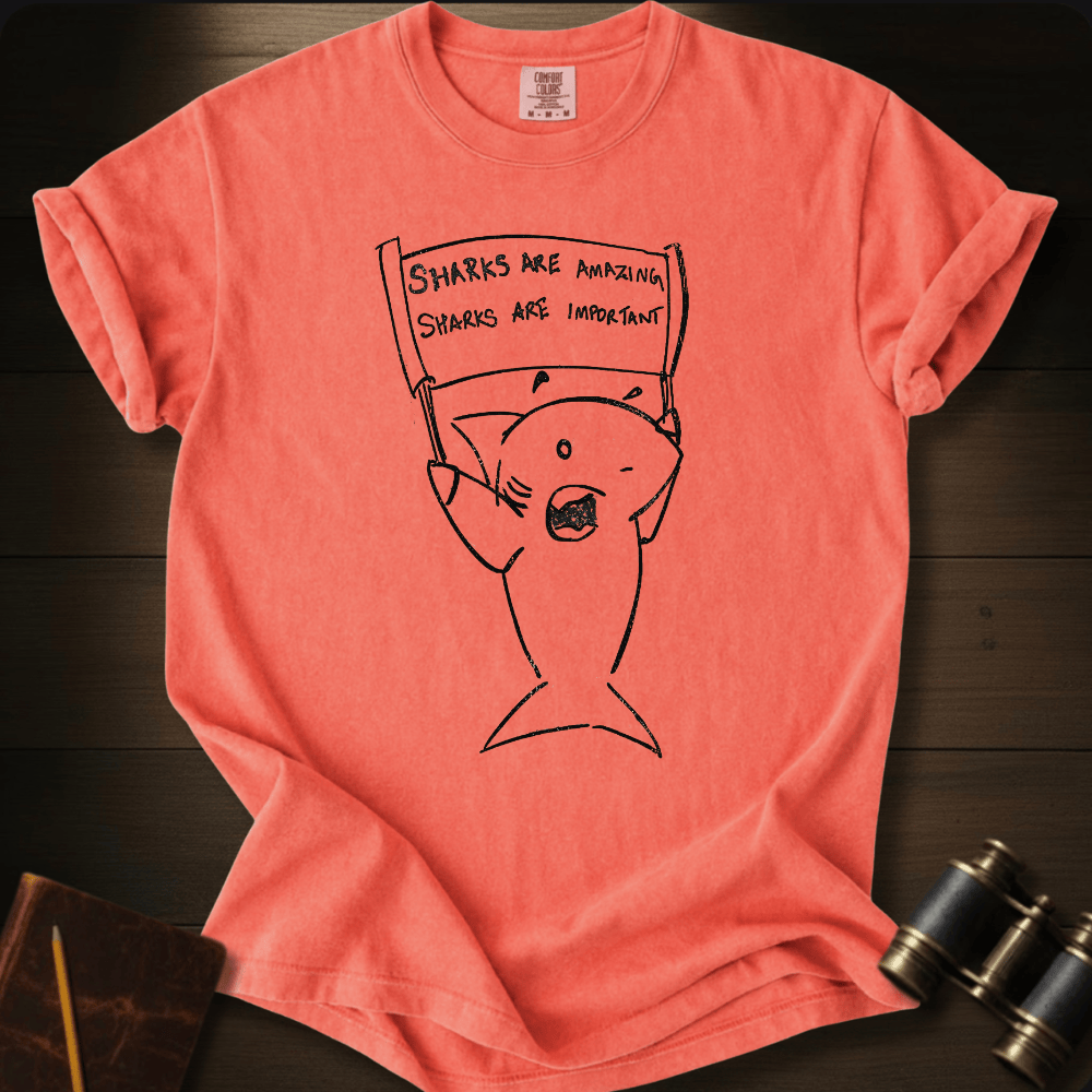 A coral pink short-sleeve t-shirt with a black line art graphic of a shark holding a protest sign that reads 'SHARKS ARE AMAZING. SHARKS ARE IMPORTANT.