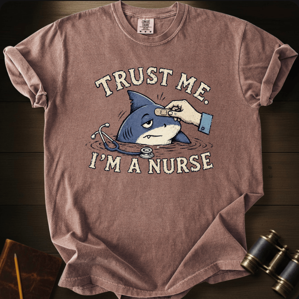 A dusty rose short-sleeve graphic t-shirt laid flat on a dark wood background. The shirt features a cartoon shark being examined by a hand with a stethoscope, with the text 'TRUST ME.