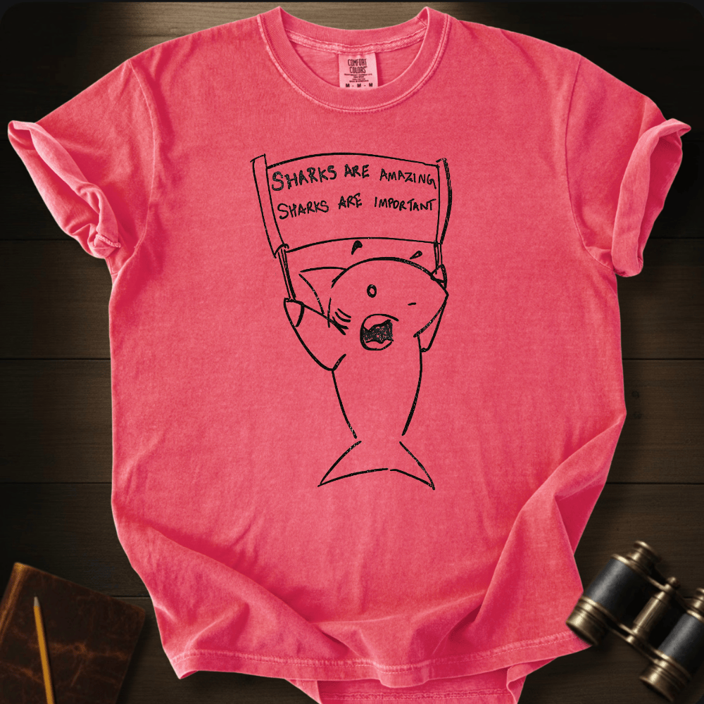 A pink short-sleeve graphic T-shirt laid flat on a dark wood surface, featuring a black line drawing of a fish holding a sign that reads 'SHARKS ARE AMAZING. SHARKS ARE IMPORTANT.