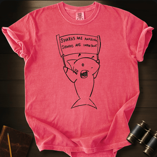 A pink short-sleeve graphic T-shirt laid flat on a dark wood surface, featuring a black line drawing of a fish holding a sign that reads 'SHARKS ARE AMAZING. SHARKS ARE IMPORTANT.