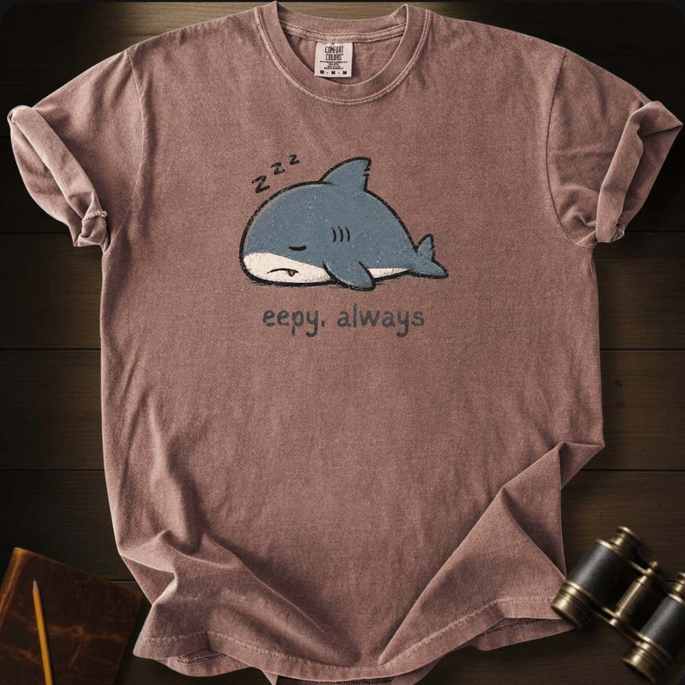 A heathered chocolate t-shirt laid flat on a dark wood background, featuring a cartoon graphic of a sleeping blue shark with 'z-z-z' above it and the text 'eepy, always' below.