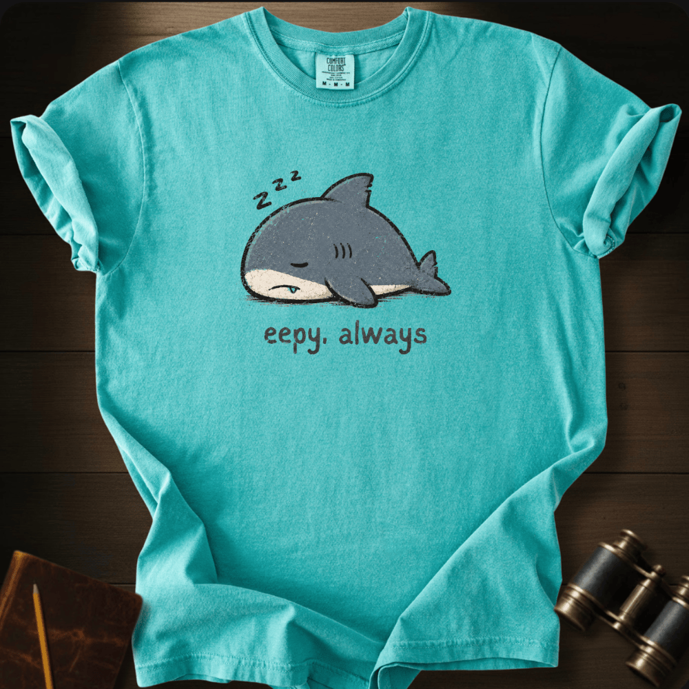 A turquoise short-sleeve crewneck t-shirt laid flat on a dark wood background, featuring a cartoon sleeping shark graphic with 'z z z' above it and the text 'eepy. always' below.