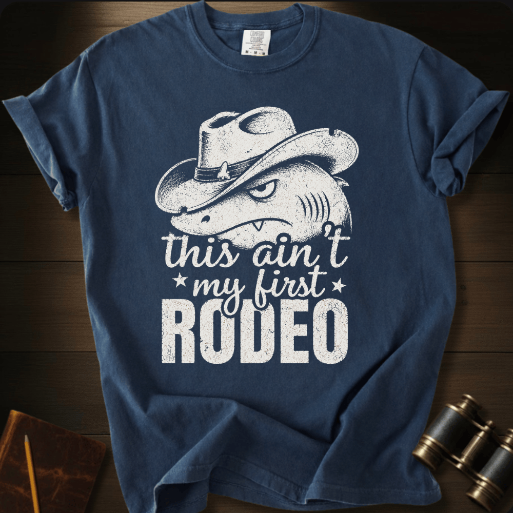 A navy blue short-sleeve t-shirt with a distressed white graphic of a shark wearing a cowboy hat and the text 'this ain't my first RODEO', displayed flat on a dark wood background with vintage binoculars and a notebook as props.