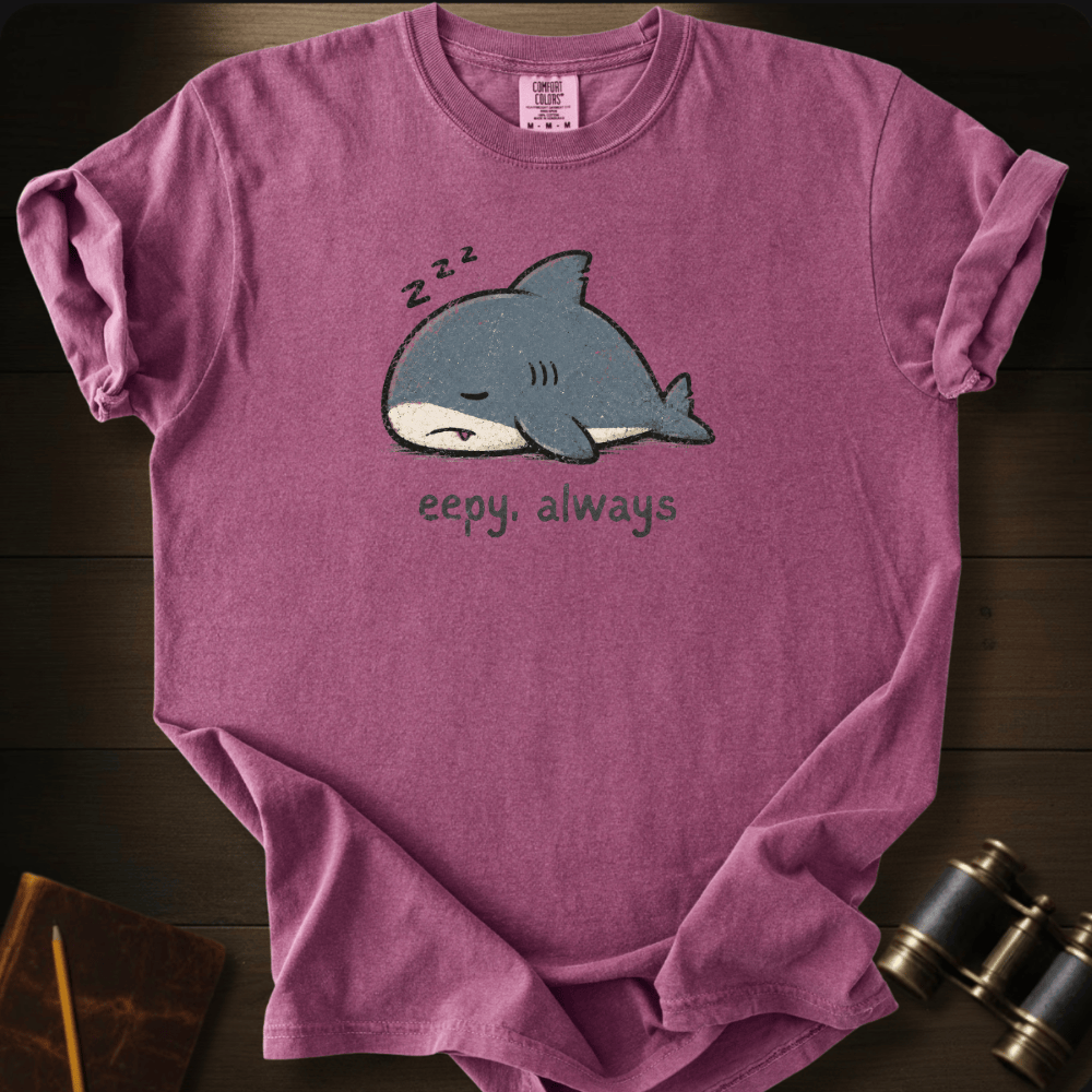 A mauve-purple short-sleeve t-shirt laid flat on a dark wood background, featuring a cartoon sleeping shark graphic with 'z-z-z' above it and the text 'eepy, always' below.