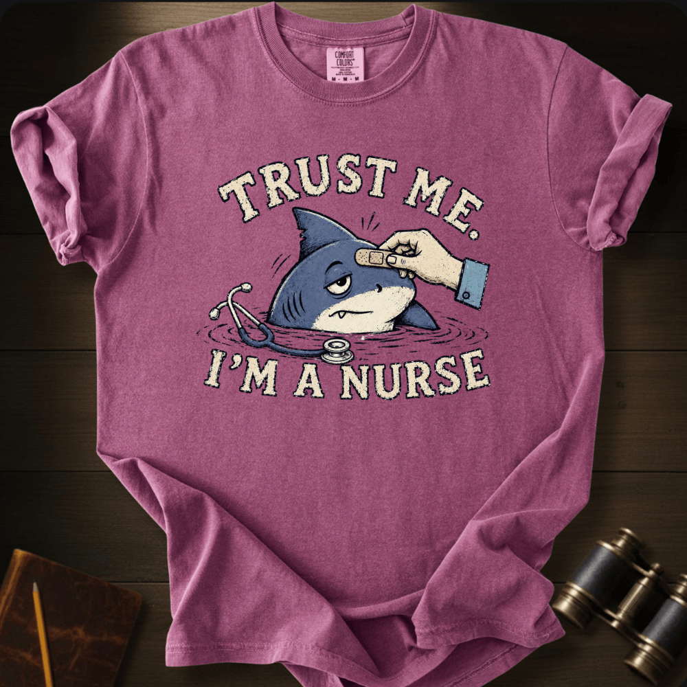 A mauve-purple short-sleeve graphic t-shirt laid flat on a dark wood background. The shirt features a cartoon shark with a stethoscope and a hand placing a bandage on its head, with the text 'TRUST ME.