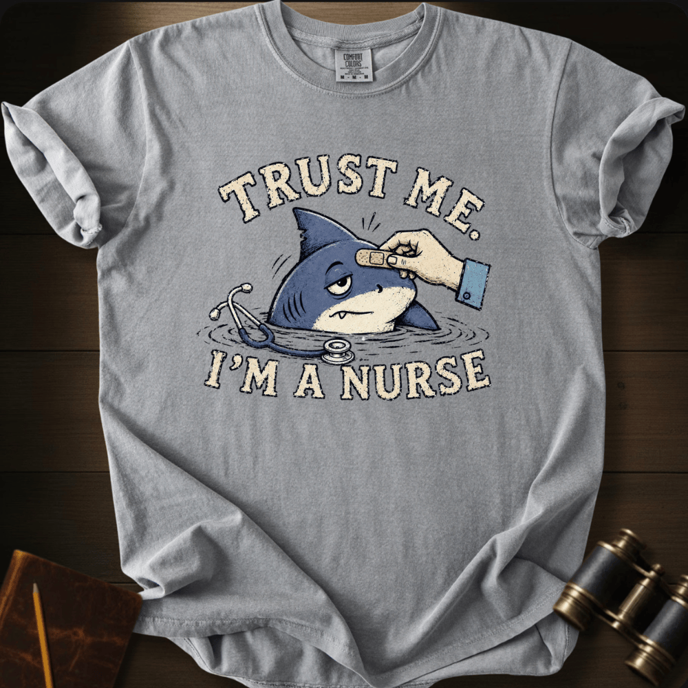 A light gray graphic t-shirt laid flat on a dark wood background. The shirt features a cartoon illustration of a shark in water with a bandage on its forehead, surrounded by the text 'TRUST ME.