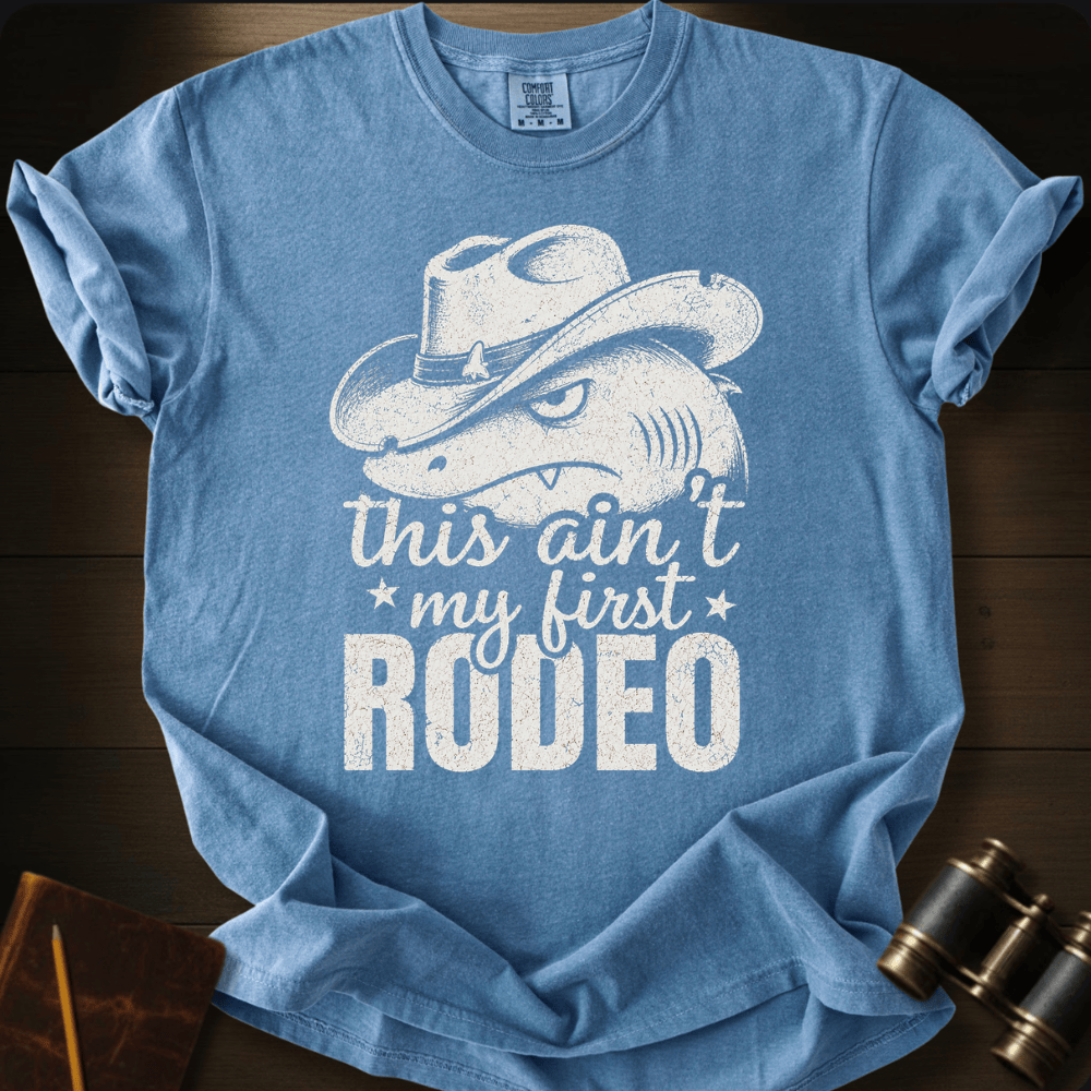 A light blue graphic t-shirt laid flat on a dark wood background, featuring a distressed white print of a shark wearing a cowboy hat with the text 'this ain't my first RODEO'.