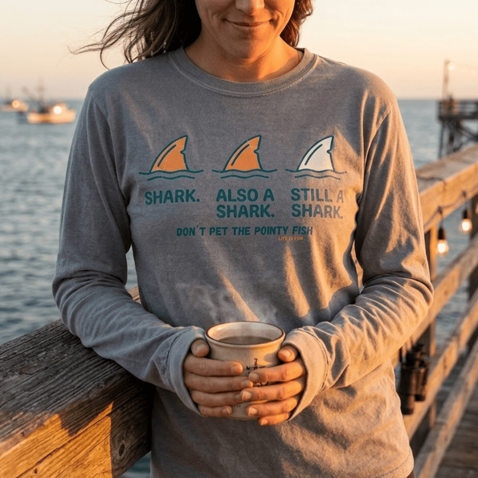 Woman wearing FullSailMarine's 'Don't Pet The Pointy Fish' shark long sleeve tee in heather grey, holding a coffee mug on a wooden pier at sunset with fishing boats in the background — ocean apparel for cooler weather